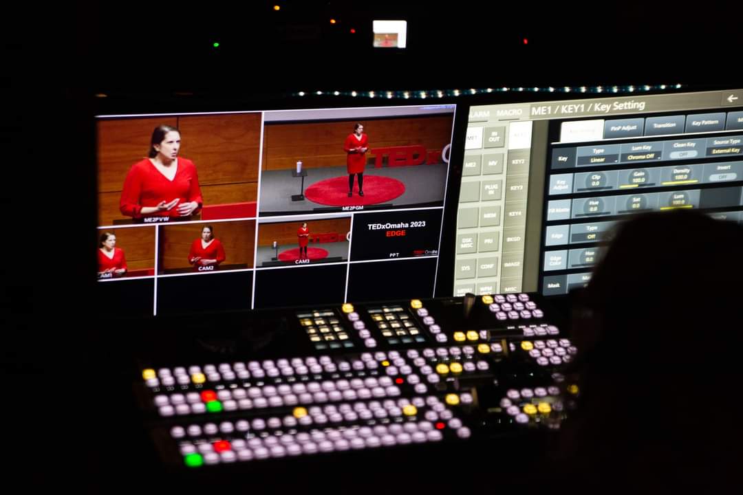 A control room monitor shows multiple camera angles of a woman in a red shirt giving a TEDx talk onstage, with a mixing console and buttons in the foreground.
