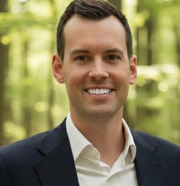 A man in a dark suit and white shirt smiles at the camera. He has short dark hair and is standing outdoors with a blurred background of green trees and sunlight.