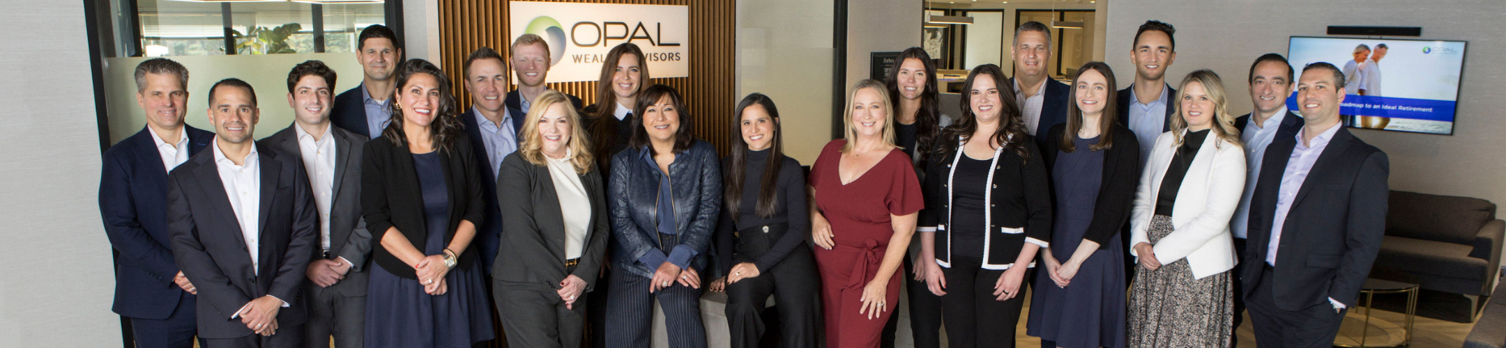 A group of 20 professionally dressed adults pose together and smile in an office lobby with a sign that reads 