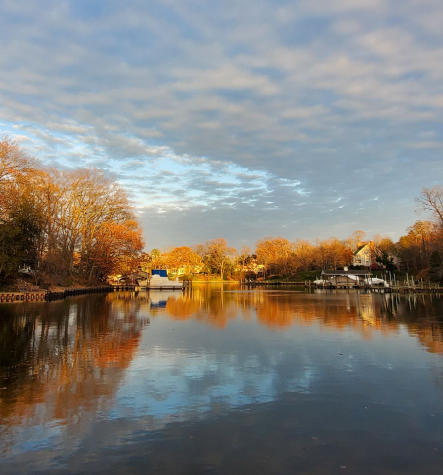 A calm lake reflects autumn trees with orange and yellow leaves, houses, and a cloudy sky at sunset. The scene is peaceful and serene, with still water mirroring the landscape.
