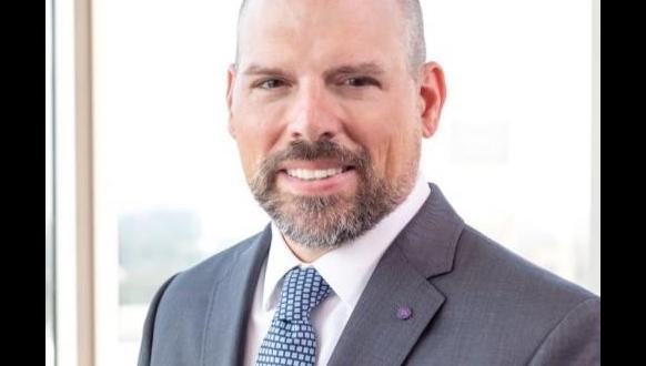 A man with a trimmed beard and shaved head is wearing a gray suit, white shirt, and patterned tie. He is smiling slightly and standing in front of a bright window with a blurred background.