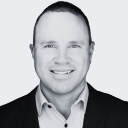 A man with short hair smiles at the camera, wearing a light-colored dress shirt and a dark blazer, in a black-and-white portrait with a plain background.