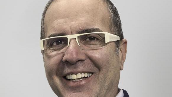 A middle-aged man with short hair and glasses smiles at the camera against a plain light gray background.