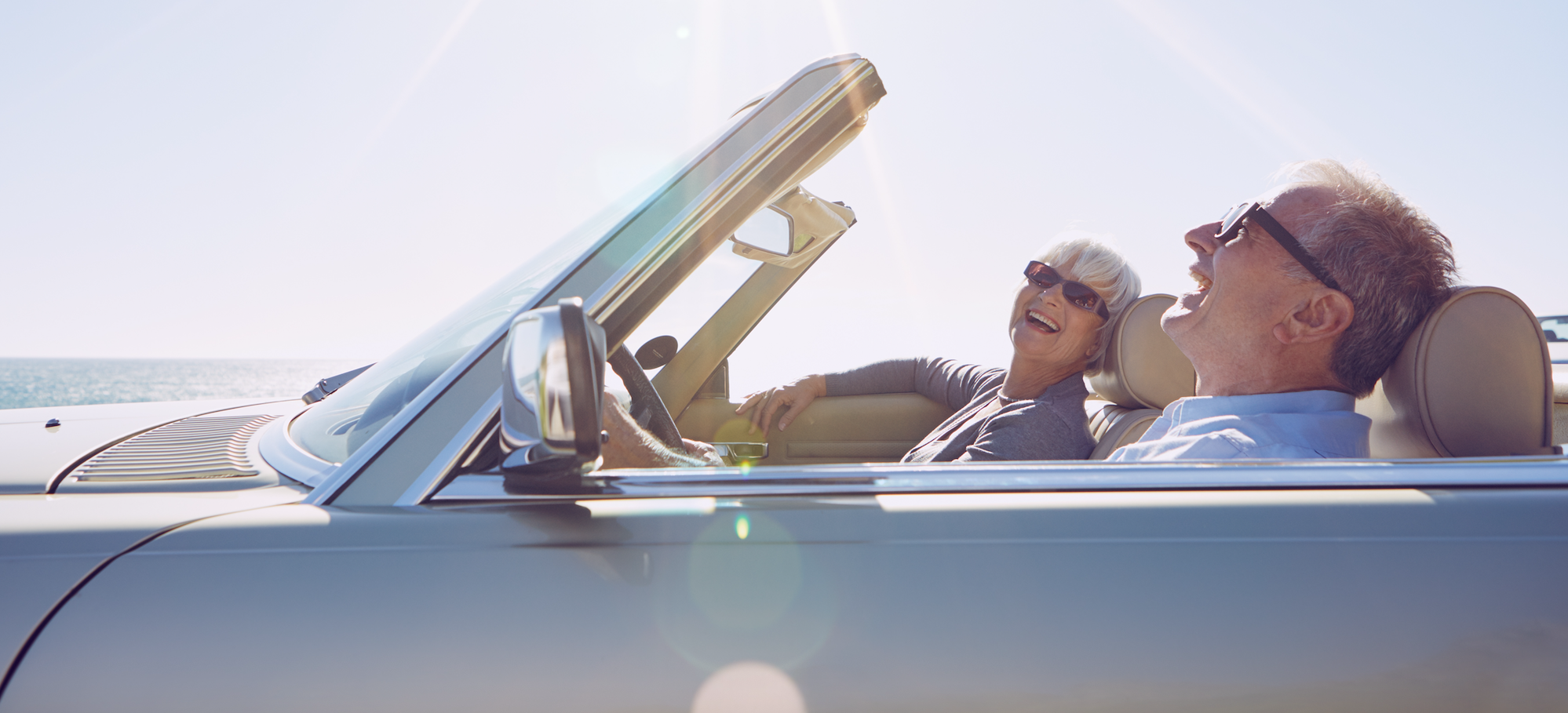 Two older adults laugh and relax in a convertible car with the top down, enjoying a sunny day by the ocean.