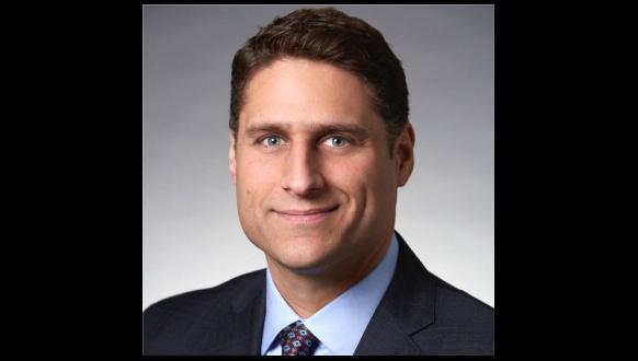 A man in a dark suit and tie, with short brown hair, smiles slightly against a plain gray background.