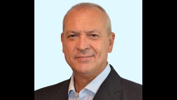 A middle-aged man with short gray hair, wearing a dark suit jacket and a light blue collared shirt, smiling softly against a light blue background.