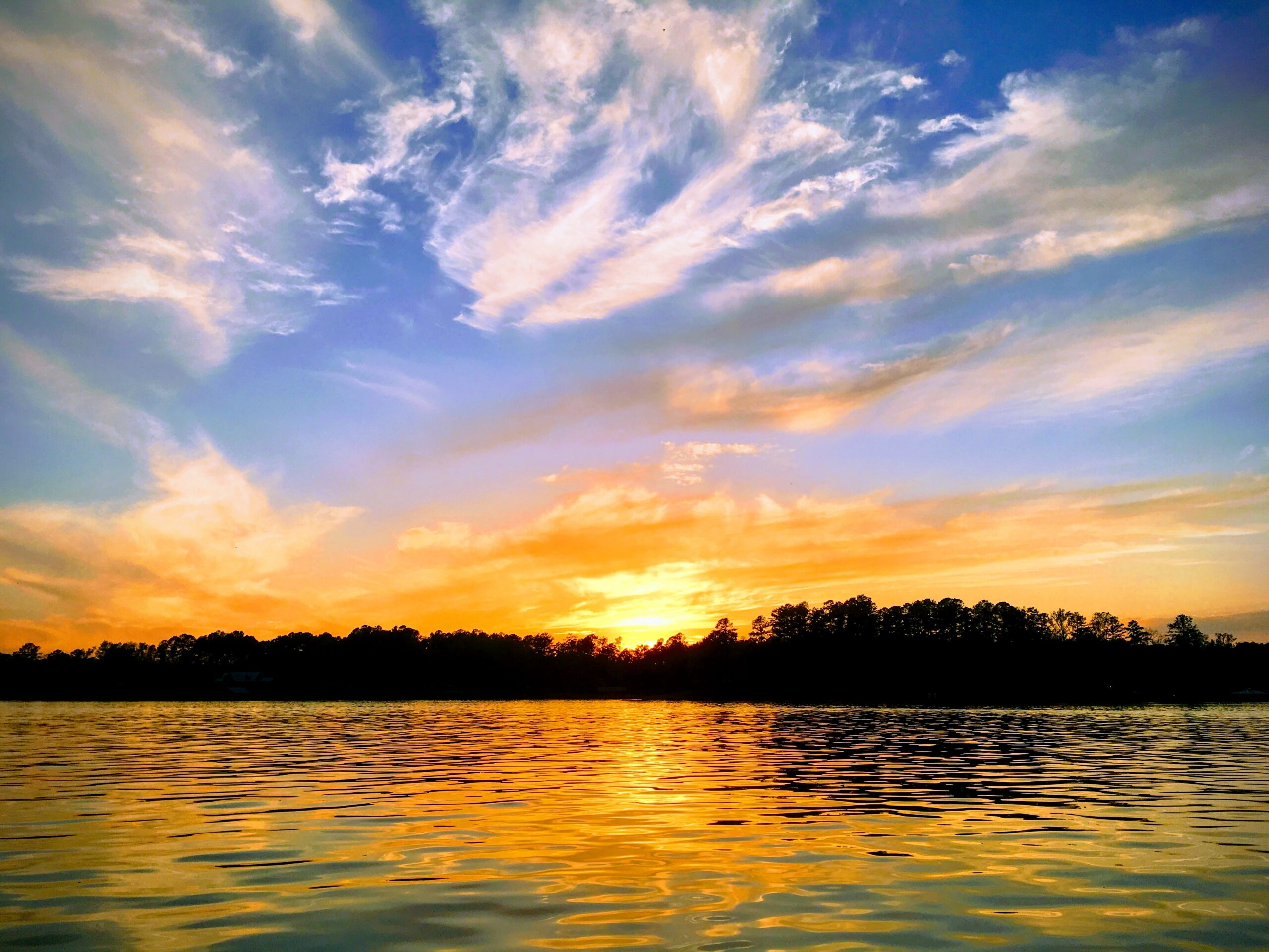 Sunset over a calm lake with a tree-lined horizon and wispy clouds in the sky.